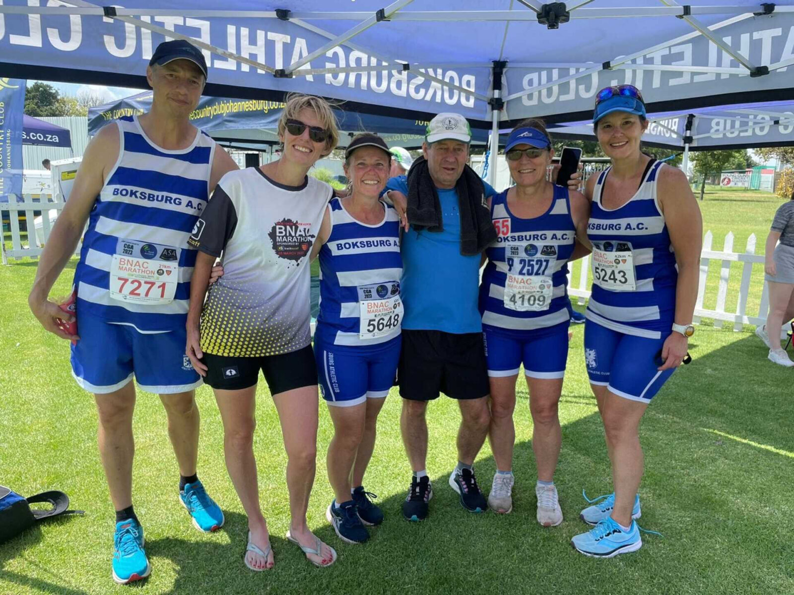 Group of Boksburg Athletic Club runners wearing official blue and white striped vests at a race event gazebo setup.