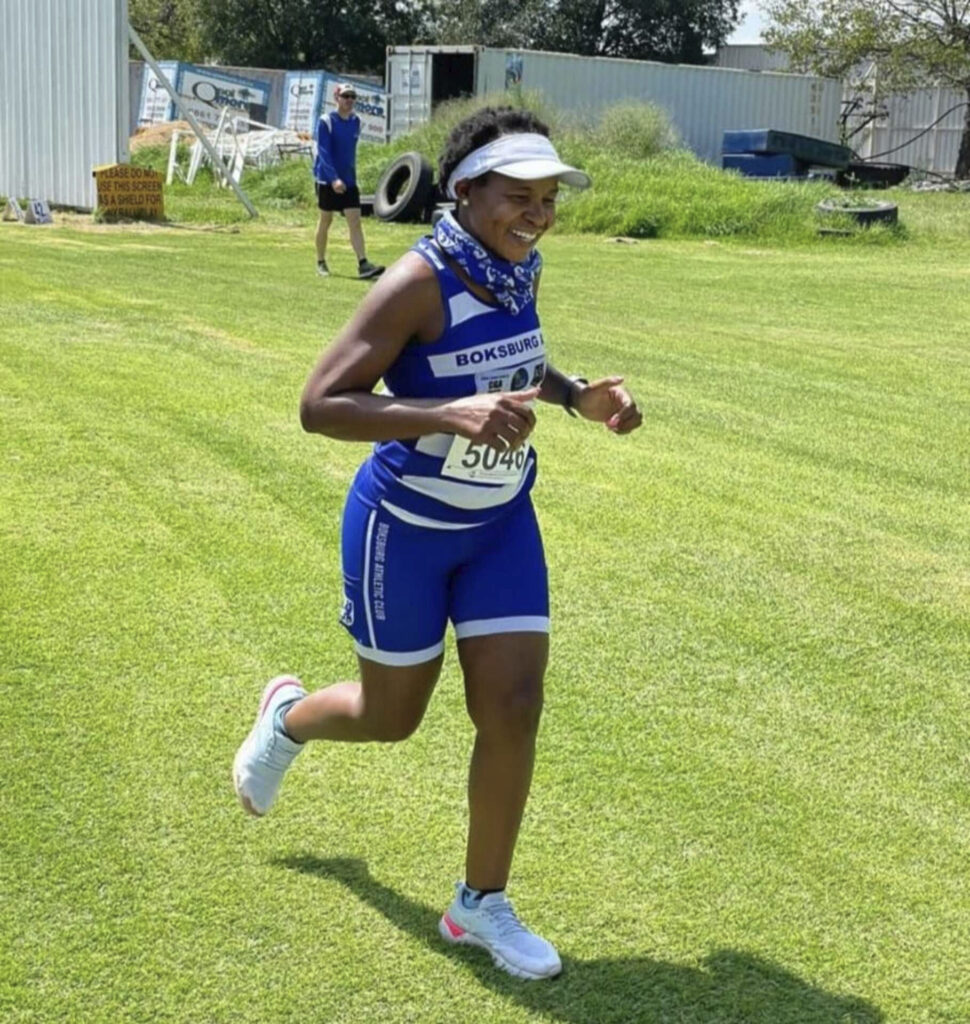 A female Boksburg Athletic Club athlete smiling while running on grass during a local club event wearing the blue and white striped running kit.
